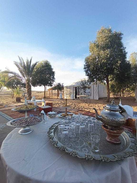 Sahara dunes Luxury Camp outdoor setup with silver tea pot, clear glasses on a tray, white tablecloth, and tents surrounded by trees