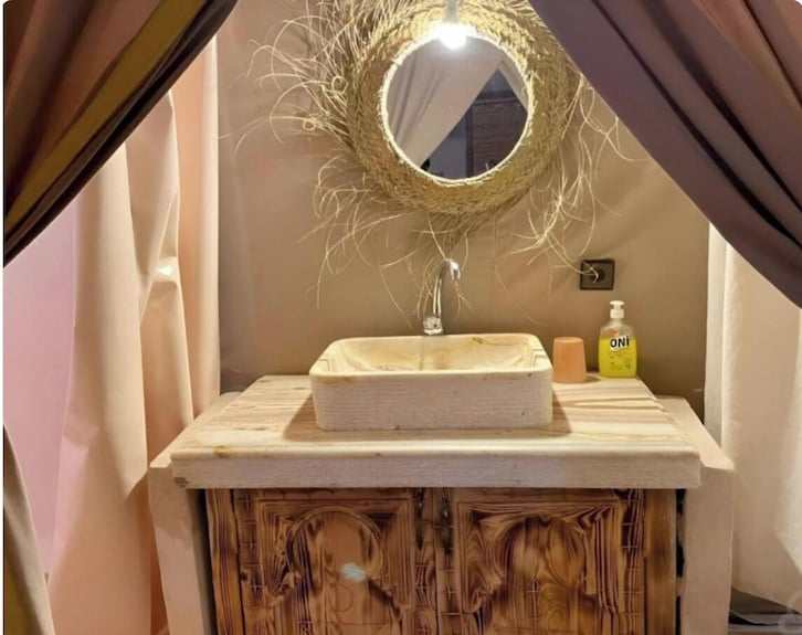 Bathroom vanity with rustic wooden cabinet, beige rectangular sink, circular straw-framed mirror, and wall-mounted faucet at Sahara dunes Luxury Camp