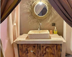 Bathroom vanity with rustic wooden cabinet, beige rectangular sink, circular straw-framed mirror, and wall-mounted faucet at Sahara dunes Luxury Camp