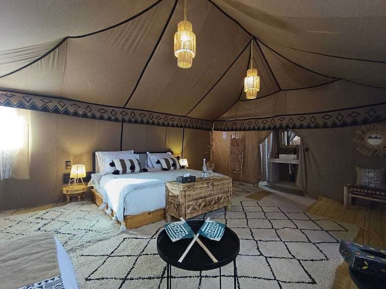 Sahara dunes Luxury Camp tent interior featuring a large bed with white linens and patterned pillows, wooden furniture, soft lighting, and a beige fabric ceiling with tribal border patterns
