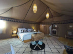 Sahara dunes Luxury Camp tent interior featuring a large bed with white linens and patterned pillows, wooden furniture, soft lighting, and a beige fabric ceiling with tribal border patterns