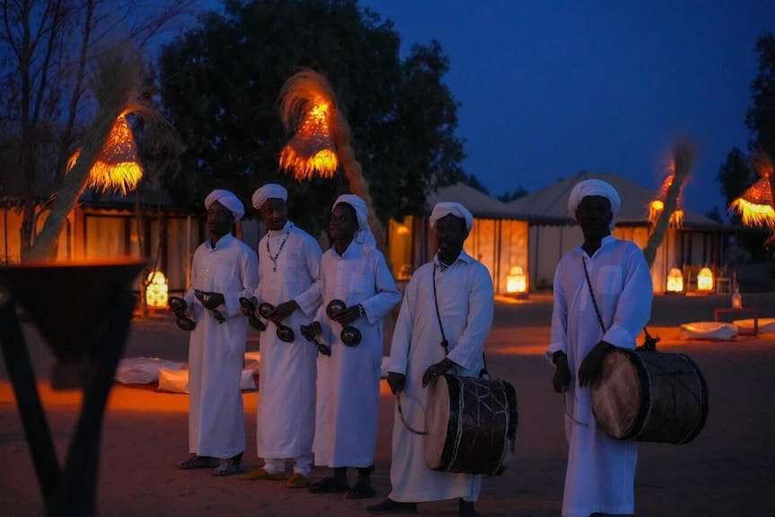 Sahara dunes Luxury Camp evening scene with five men in traditional white attire playing musical instruments under warm lantern light