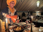 Sahara dunes Luxury Camp man in traditional clothing serving a tagine dish inside an ornate tented dining area with candles and tableware