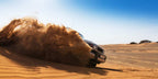 White off-road vehicle kicking up a large cloud of sand in a desert with rippled dunes under blue sky, Sahara dunes Luxury Camp