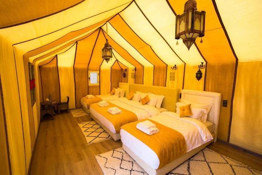 Sahara dunes Luxury Camp spacious tent interior with yellow and cream canvas walls, three beds with orange throws and hanging lanterns