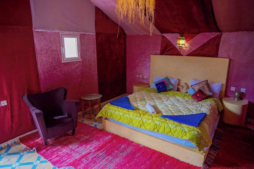 Sahara Garden Luxury Camp bedroom featuring a large bed with colorful pillows, yellow quilt, purple armchair, and red carpet