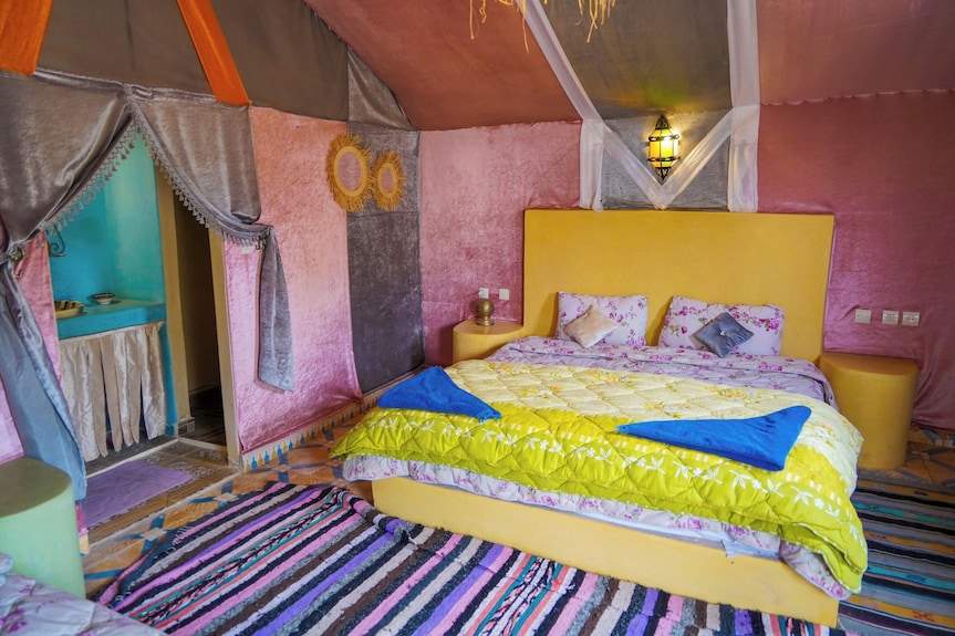 Sahara Garden Luxury Camp bedroom with yellow bed, floral sheets, pink and purple walls, colorful striped rugs, and Moroccan style decor