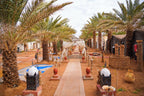 Sahara Garden Luxury Camp desert walkway lined with palm trees, traditional Berber pottery, lanterns, and tents in warm sandy tones