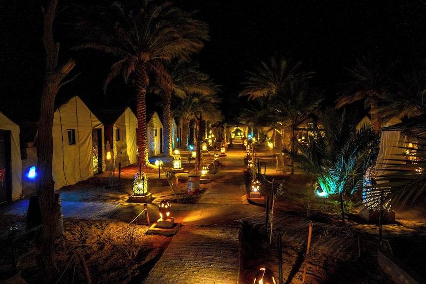 Sahara Garden Luxury Camp night view with illuminated desert tents, palm trees, and lantern-lined pathway in warm lighting