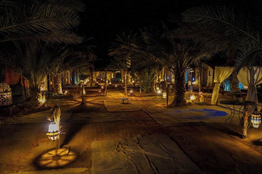 Sahara Garden Luxury Camp outdoor night scene with lit lanterns, palm trees, and sandy ground pathways under a dark sky