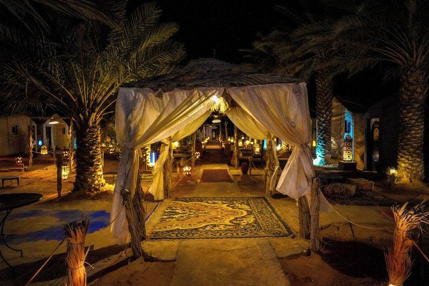 Sahara Garden Luxury Camp nighttime entrance with illuminated white fabric canopy, patterned carpets, and palm trees