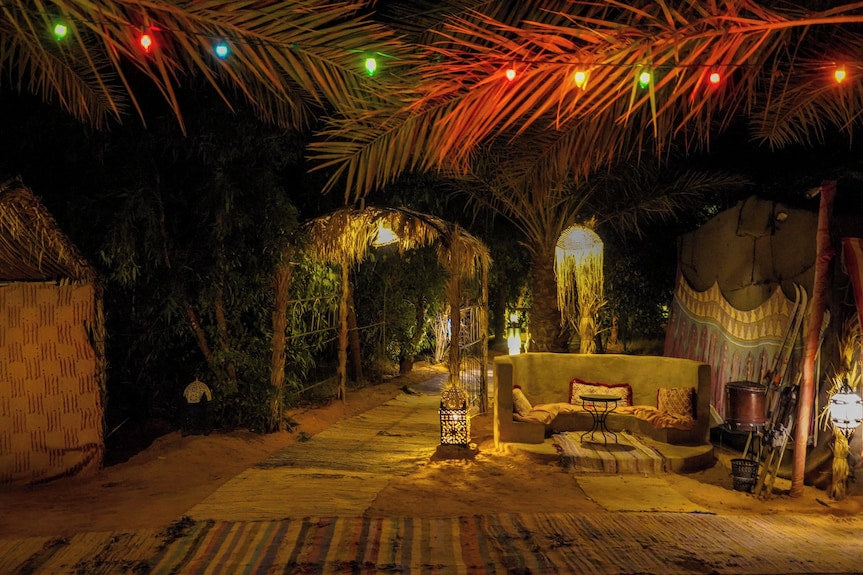 Sahara Garden Luxury Camp outdoor seating area at night with warm lighting, colorful string lights, palm trees, and traditional Moroccan decor