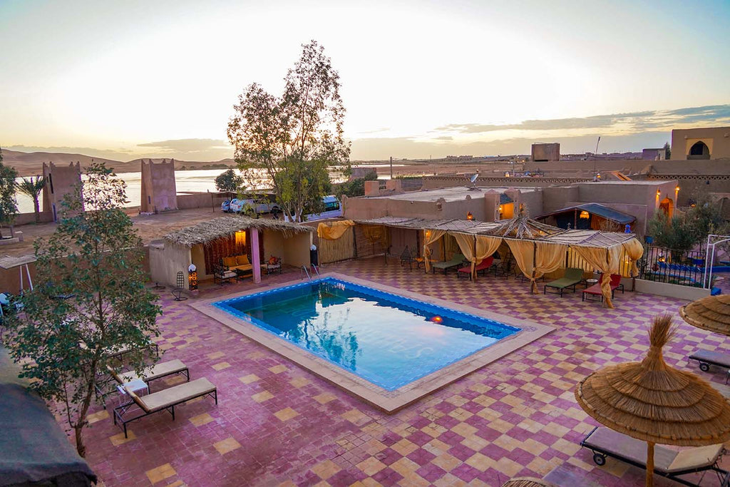 Sahara Garden Luxury Camp outdoor area with a rectangular blue swimming pool, tiled courtyard, shaded seating areas with beige curtains, and surrounding desert landscape at sunset