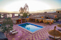 Sahara Garden Luxury Camp outdoor area with a rectangular blue swimming pool, tiled courtyard, shaded seating areas with beige curtains, and surrounding desert landscape at sunset