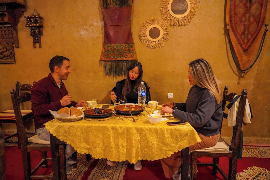 Dining scene inside Sahara Garden Luxury Camp with three people eating traditional Moroccan meal at round table with yellow tablecloth