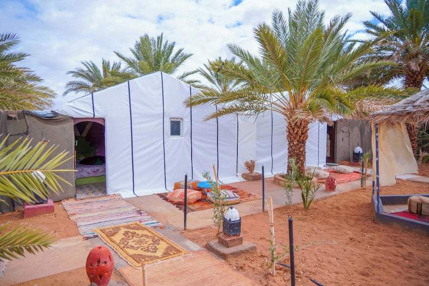 Sahara Garden Luxury Camp with large white tents, colorful rugs, cushions, palm trees, and sandy courtyard in desert garden