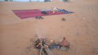 Sahara Trek Camp setup with a small open fire, traditional clay tagine pot, and a red textured rug with tea set on sandy desert ground