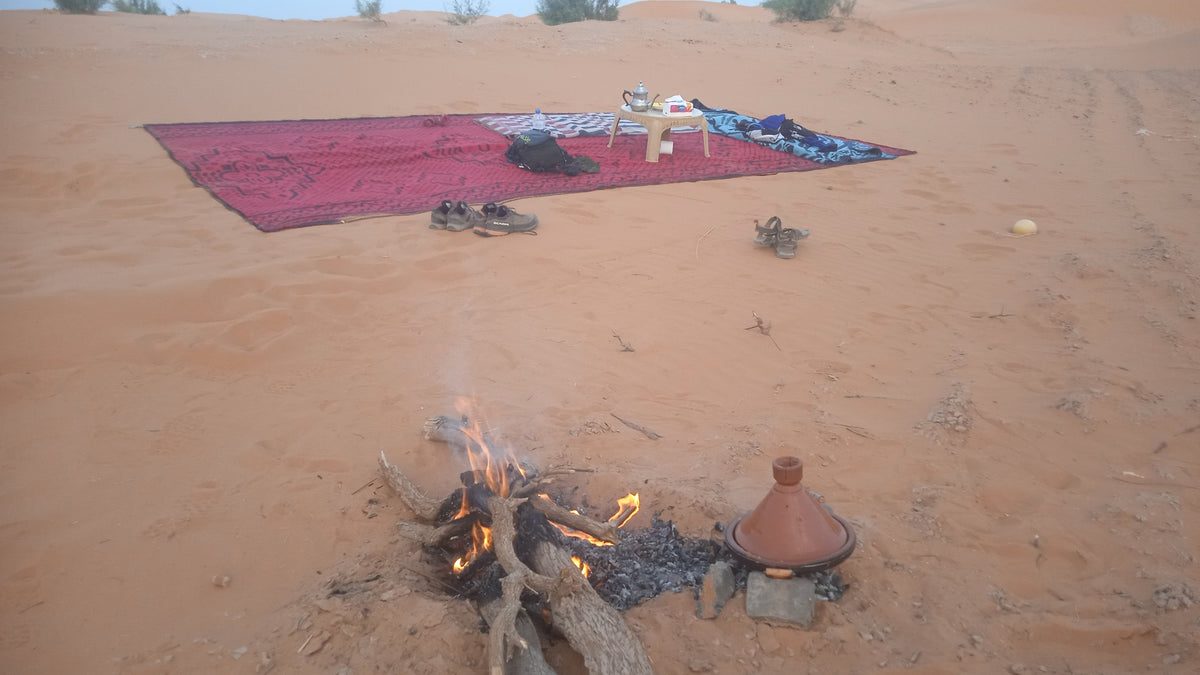 Sahara Trek Camp setup with a small open fire, traditional clay tagine pot, and a red textured rug with tea set on sandy desert ground