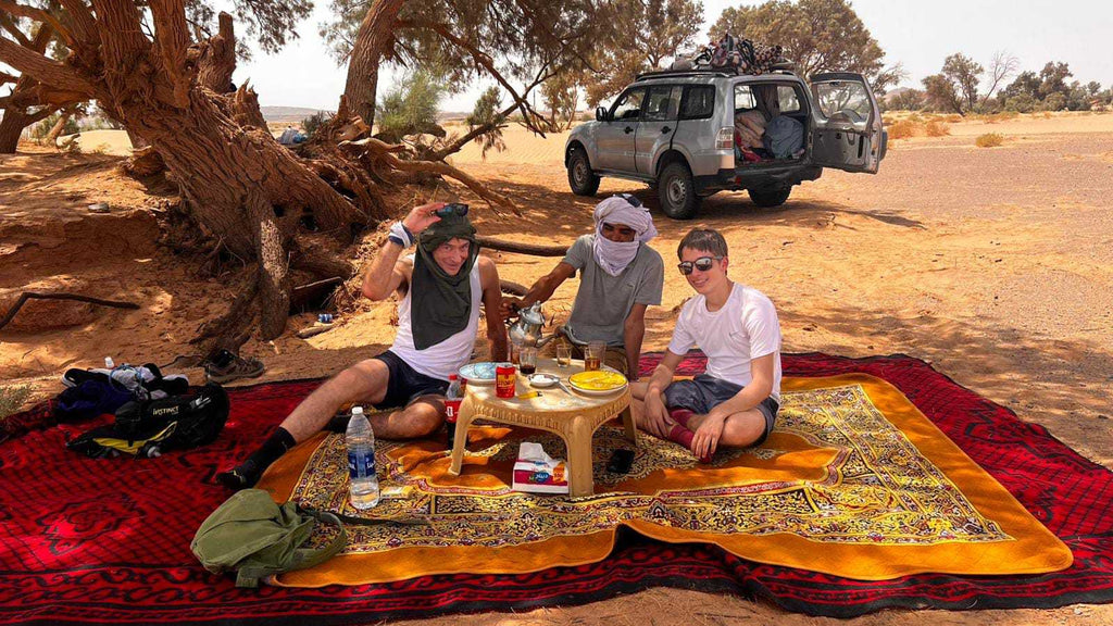 Outdoor picnic setup with colorful layered rugs and a small table, featuring people enjoying traditional tea near a Sahara Trek Camp.