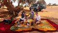 Outdoor picnic setup with colorful layered rugs and a small table, featuring people enjoying traditional tea near a Sahara Trek Camp.