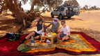 Outdoor picnic setup with colorful layered rugs and a small table, featuring people enjoying traditional tea near a Sahara Trek Camp.
