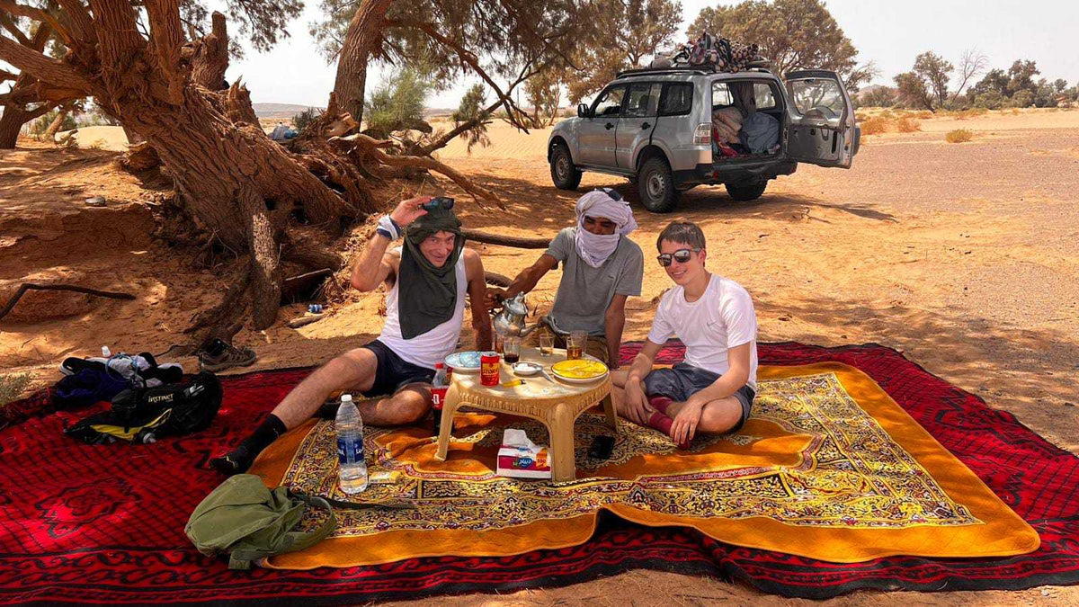 Outdoor picnic setup with colorful layered rugs and a small table, featuring people enjoying traditional tea near a Sahara Trek Camp.