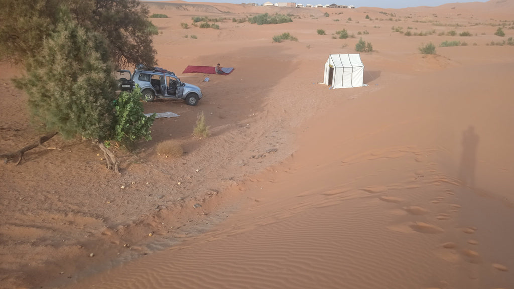 Sahara Trek Camp showing a white canvas tent and silver 4x4 vehicle parked near sparse greenery in sandy desert dunes under clear sky