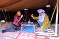 Sahara Wild Camp interior with two people sitting on patterned rugs under a shaded brown tent, enjoying drinks at a low round table.