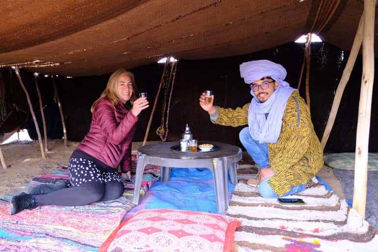 Sahara Wild Camp interior with two people sitting on patterned rugs under a shaded brown tent, enjoying drinks at a low round table.