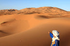 Sahara Wild Camp desert landscape with vast golden sand dunes and a person in a blue robe and white headwrap pointing
