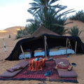 Desert tent with brown fabric, beds inside with white and blue linens, cushions, and rugs outside on sand at Standard Camp Merzouga Oasis