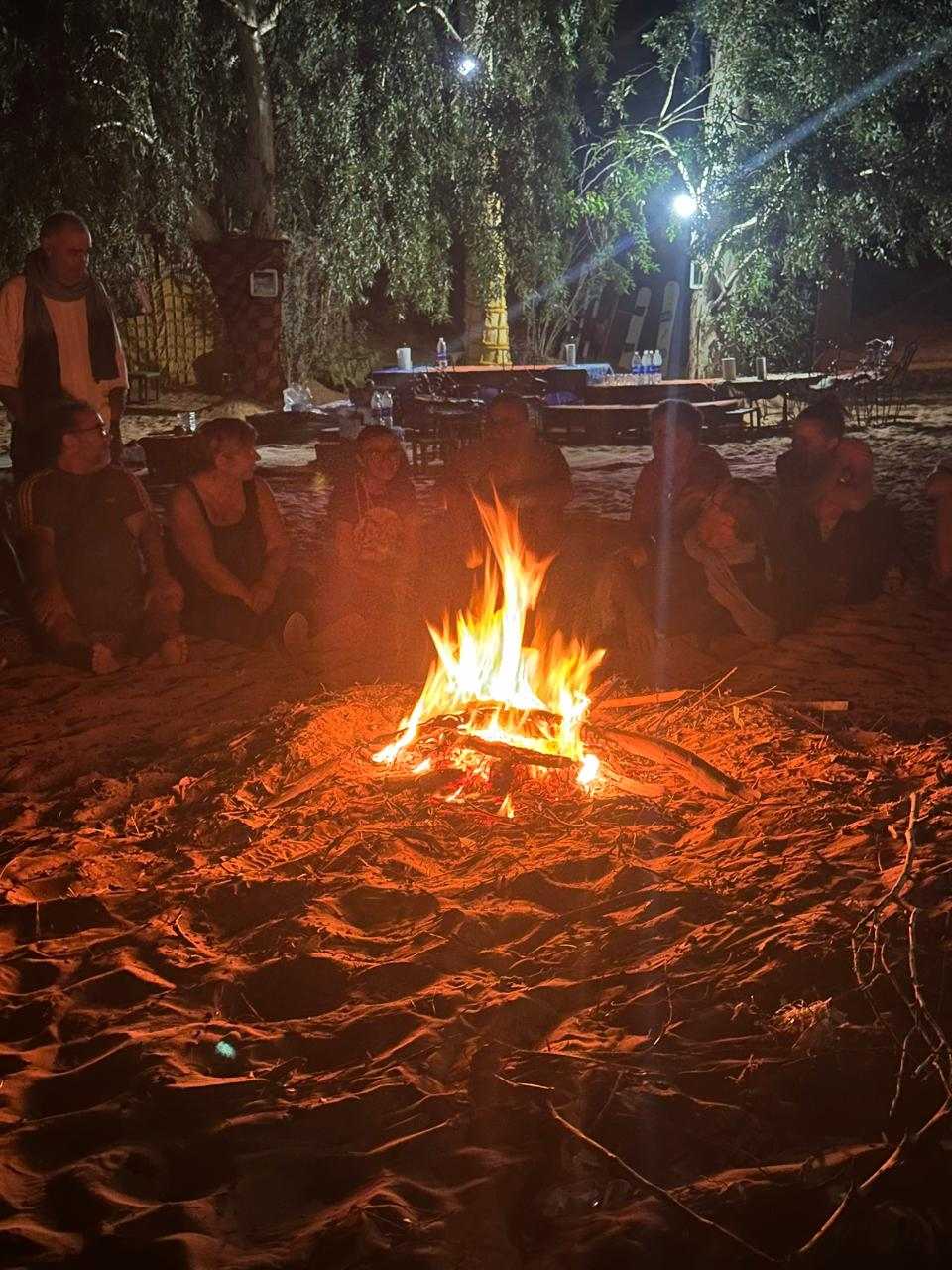 Sahara Wild Camp at night with a bright campfire surrounded by people sitting on sand under trees