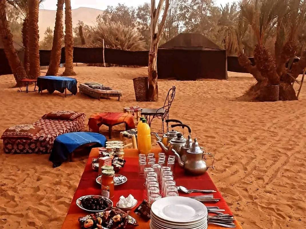 Sahara Wild Camp outdoor dining setup on red sand with traditional cushions, metal teapots, glasses, and plates under palm trees