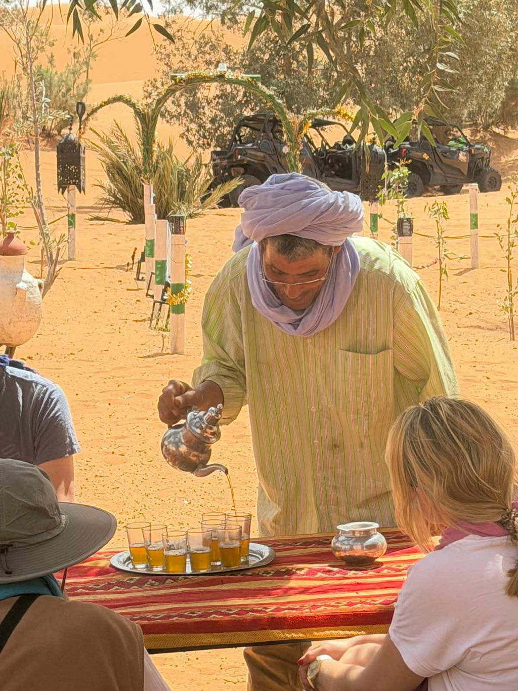 Sahara Wild Camp with person in light green striped shirt and purple headscarf pouring tea into small glasses on red patterned tablecloth outdoors