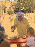 Sahara Wild Camp with person in light green striped shirt and purple headscarf pouring tea into small glasses on red patterned tablecloth outdoors