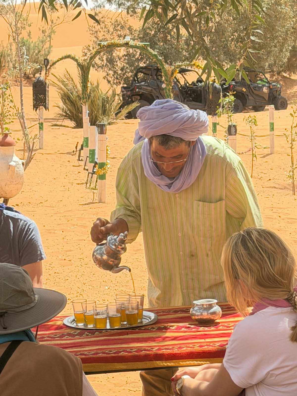 Sahara Wild Camp with person in light green striped shirt and purple headscarf pouring tea into small glasses on red patterned tablecloth outdoors