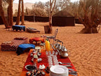 Sahara Wild Camp outdoor setup with traditional low cushions, red tablecloth, metal tea pots, glasses, and plates on desert sand