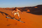 Young person sandboarding on orange sand dunes under a clear blue sky illustrating Sandboarding and Watching the Sunset adventure