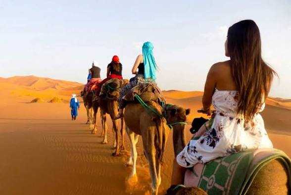 Camel caravan with travelers wearing colorful clothing riding through golden sand dunes during a shared 3 days 2 nights desert tour.