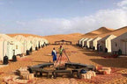 Desert camp with white canvas tents, sandy ground, wooden seating area, and people under clear sky during shared 3 days 2 nights tour