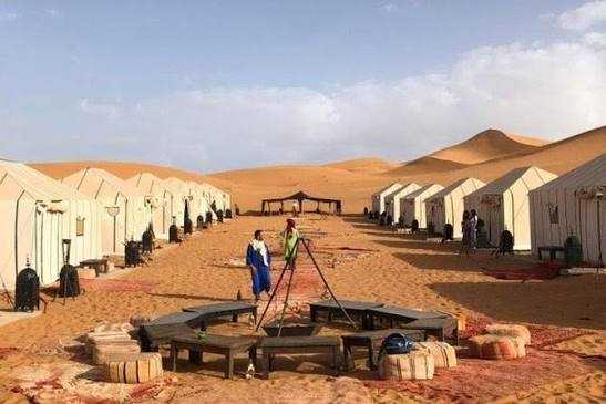 Desert camp with white canvas tents, sandy ground, wooden seating area, and people under clear sky during shared 3 days 2 nights tour