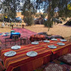 Standard Camp Merzouga Oasis outdoor dining setup with low table covered by red striped cloth, white plates, and cushions on sandy desert ground