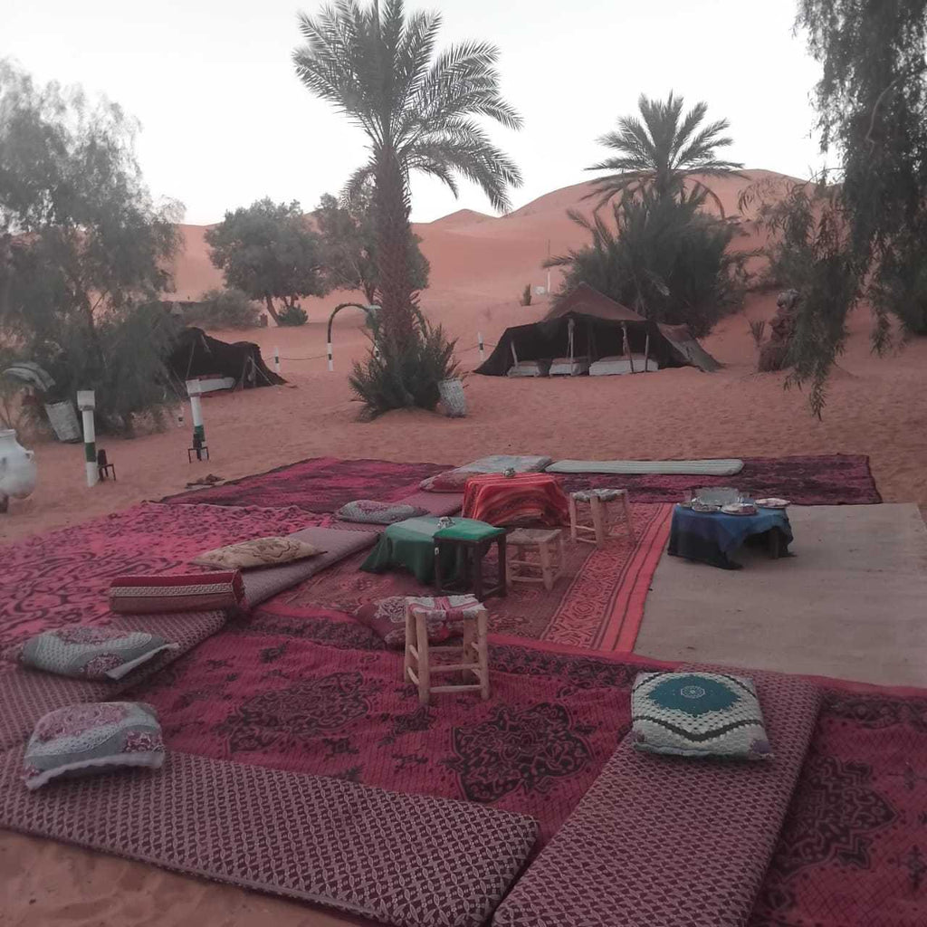 Standard Camp Merzouga Oasis outdoor seating area with Moroccan patterned rugs, cushions, small stools, palm trees, and desert tents