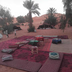 Standard Camp Merzouga Oasis outdoor seating area with Moroccan patterned rugs, cushions, small stools, palm trees, and desert tents