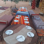Standard Camp Merzouga Oasis outdoor seating with patterned cushions and carpets around tables set with white plates and cutlery on sandy ground