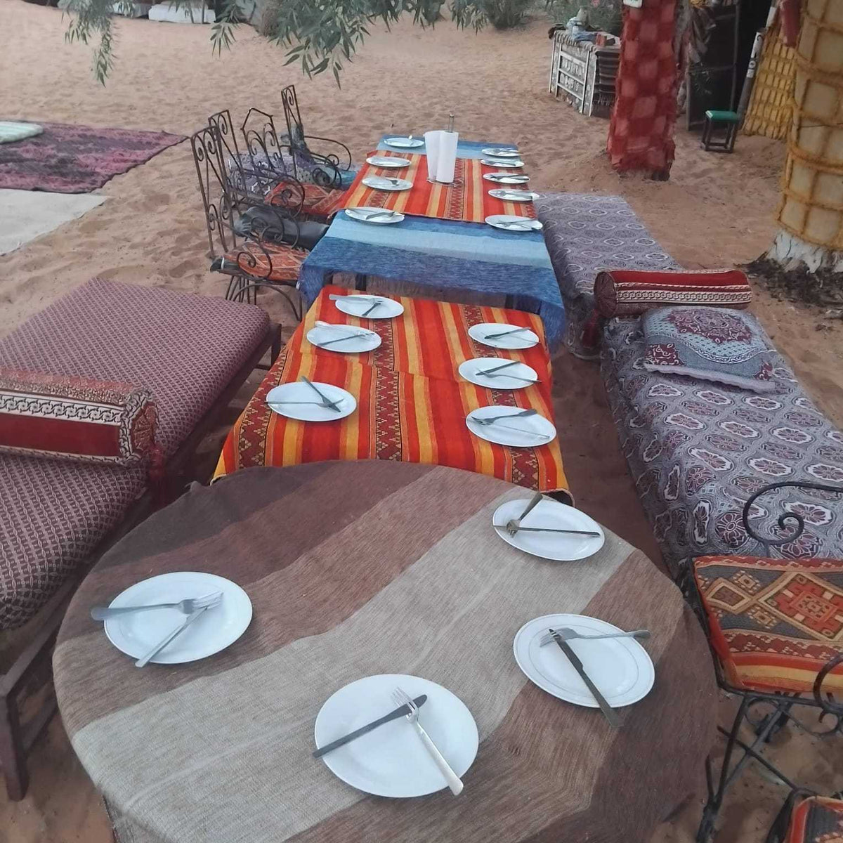 Standard Camp Merzouga Oasis outdoor seating with patterned cushions and carpets around tables set with white plates and cutlery on sandy ground