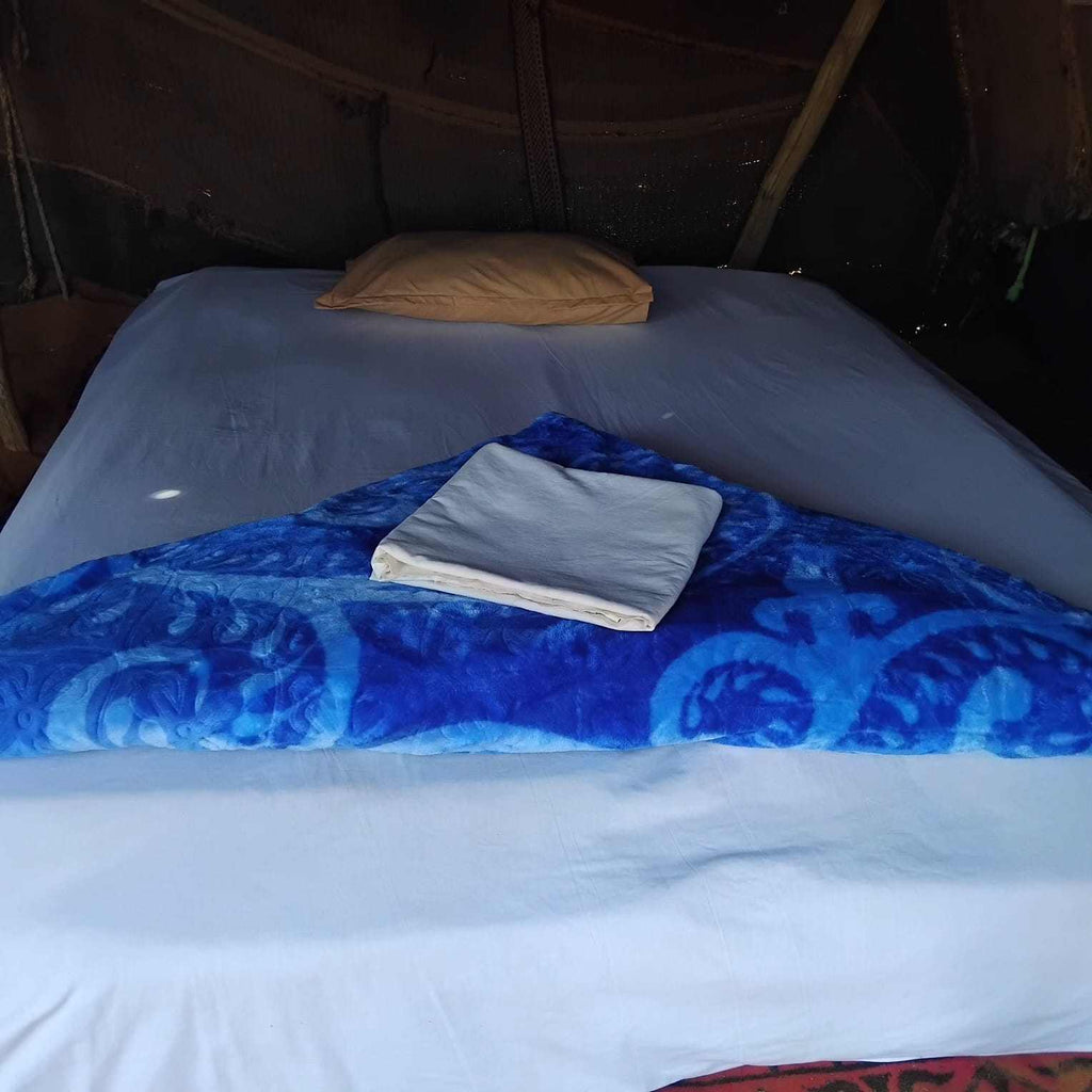 Standard Camp Merzouga Oasis tent bed with white sheets, beige pillow, and blue patterned blanket for desert comfort