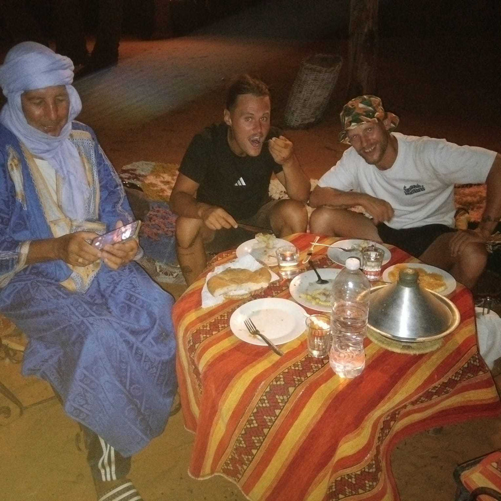 Standard Camp Merzouga Oasis featuring three people sitting around a small striped red and orange table with traditional Moroccan dishes and candles at night