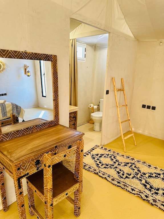 Sunset Desert Camps interior showing beige canvas walls, patterned wooden dressing table and chair, a rustic wooden ladder, and a navy patterned rug on yellow floor