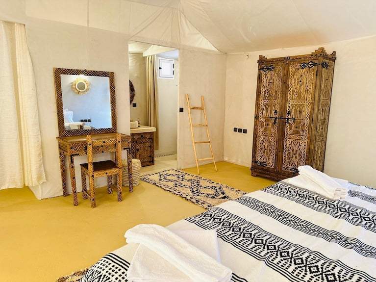 Desert camp interior at Sunset Desert Camps with beige canvas walls, yellow floor, patterned bedspread, and carved wooden furniture.
